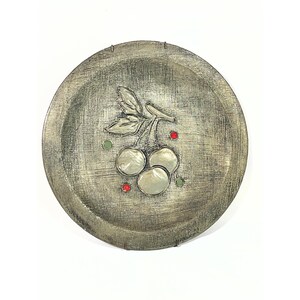 May include: A silver-colored decorative plate with a textured surface. The plate features a raised design of cherries with a leaf and stem. The cherries are silver with red and green accents.