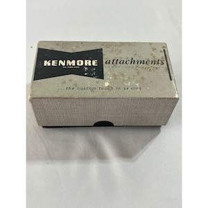 Puede incluir: Caja de accesorios Kenmore vintage. La caja es rectangular con un patrón de cuadros blancos y negros en la parte inferior. La parte superior de la caja es de color gris claro con el logotipo de Kenmore y la palabra "attachments" impresa. El texto "...the custom touch in sewing" también está impreso en la caja.