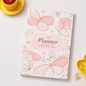 Könnte beinhalten: Ein weißer 2026-Planer mit rosa Schmetterlingen und floralen Designs. Der Planer hat das Wort "Planner" in einem rosa Rahmen, mit dem Text "A start to something new". Eine gelbe Schale mit einer kleinen Tasse und einem Lineal befindet sich links.