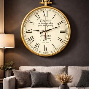 Puede incluir: Un gran reloj de pared con marco dorado, números romanos y manecillas negras. La esfera es de color crema con el texto "Every second is precious when spent with family." y "Johnson's EST 1916". El reloj está sobre un sofá gris.