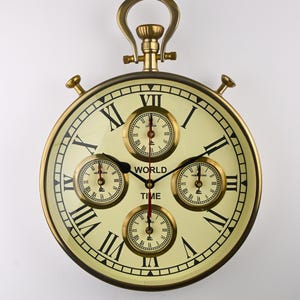 Puede incluir: Un reloj de pared redondo de estilo antiguo con esfera crema y números romanos negros. Cuenta con cuatro esferas más pequeñas y un marco y asa de color latón. La palabra "WORLD TIME" está impresa en la esfera.