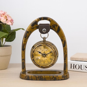 May include: A vintage-style clock with a gold-colored frame and a round clock face. The clock is suspended within an arched stand, featuring a brown leather strap at the top. The clock face has Roman numerals and decorative hands. The base is rectangular.