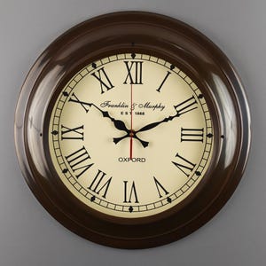 May include: A round brown wall clock with a cream-colored face. The clock features black Roman numerals, black hands, and the text "Franklin & Murphy Est 1868" and "OXFORD". The clock has a dark brown frame.