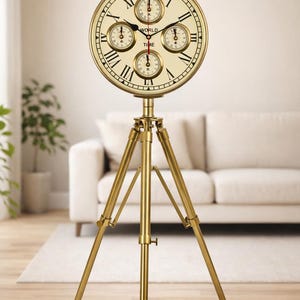 May include: A gold-colored world time clock on a tripod stand. The clock face is cream-colored with black Roman numerals and smaller dials. The words "WORLD TIME" are printed on the clock face. The tripod has three legs.