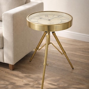 May include: A round side table with a clock face design. The table has a gold-colored metal frame and a glass top. The table is supported by three gold-colored legs. The clock face has Roman numerals and the text "Paris, France".