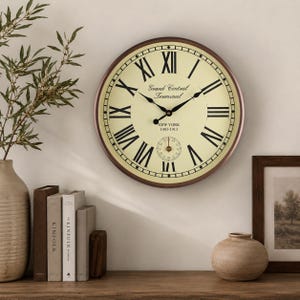 Puede incluir: Un reloj de pared redondo con marco marrón oscuro y esfera color crema con números romanos negros. El centro muestra el texto Grand Central Terminal, NEW YORK, y 1903-1913, con manecillas negras y un pequeño dial secundario en la parte inferior.