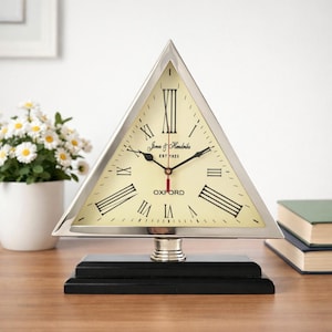 May include: A triangular clock with a silver frame and a cream-coloured face. The clock features black Roman numerals and the text "Jones & Hendricks EST 1923 OXFORD". The clock sits on a black base.
