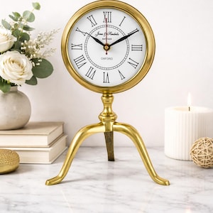 May include: A gold-colored, tripod-based clock with a white face and black Roman numerals. The clock face has the text "Jones & Hendrikx, EST 1923" and "OXFORD". The clock is displayed on a marble surface, with a candle and decorative items in the background.
