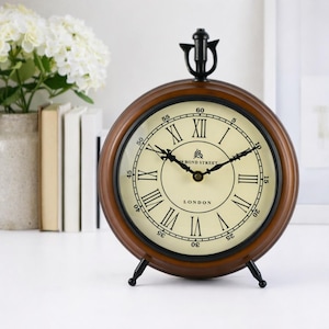 Puede incluir: Un reloj redondo de madera marrón con esfera crema y números romanos negros. El reloj tiene manecillas negras y una base de metal negro. El texto "19 BOND STREET LONDON" está impreso. Un artículo decorativo para el hogar.