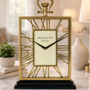 Puede incluir: Un reloj rectangular dorado con números romanos y una base negra. La esfera del reloj es de color crema con manecillas negras y el texto "McLaughlin & Scott ESTD 1972". El reloj tiene un asa decorativa en la parte superior.