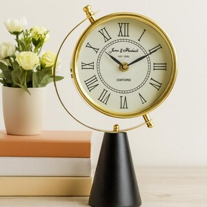 May include: A gold-coloured desk clock with a black conical base. The clock face is cream with black Roman numerals and hands. The clock is labelled "James & Mansfield, EST 1883, Oxford". The word "HOME" is in the background.