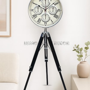 May include: A large, round clock with multiple dials, mounted on a black tripod stand. The clock face features Roman numerals and smaller dials. The stand has black legs and silver metal accents. The text "LUMO CRAFT DECOR" is visible.