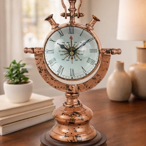 May include: A copper-colored decorative clock with a compass rose design. The clock features Roman numerals and a glass face. The clock sits on a wooden base and has a unique, vintage aesthetic. The clock is a decorative home decor item.