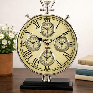 Puede incluir: Un reloj de hora mundial plateado con una esfera color crema y números romanos negros. El reloj presenta cuatro esferas más pequeñas y las palabras "WORLD TIME". Se asienta sobre una base negra.