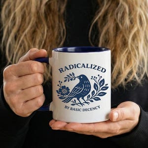 May include: White ceramic mug with a blue interior and handle. The mug features the word "RADICALIZED" above a blue illustration of a bird with a floral and leaf design. The text "BY BASIC DECENCY" is below the bird.