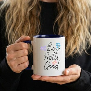 May include: A white ceramic mug with a blue interior and handle. The mug displays the text "Be Pretti Good" in a script font, accompanied by purple and blue floral and butterfly illustrations. The mug is held by a person.