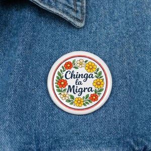 Puede incluir: Un botón redondo blanco con el texto "Chinga la Migra" en una fuente estilizada, rodeado por una corona de flores coloridas. El botón está prendido a una chaqueta vaquera.