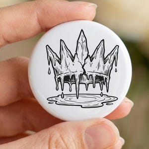 May include: A white button with a black and white illustration of a crown with a cracked center and dripping details. The button is held in a hand, with a blurred floral background.