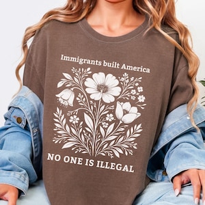 Puede incluir: Camiseta color espresso con un diseño floral blanco y el texto "Immigrants built America" y "NO ONE IS ILLEGAL." Un estilo casual y cómodo.