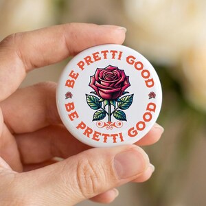 Peut inclure: Un bouton blanc avec les mots "BE PRETTI GOOD" en orange sur le bord. Une rose rouge avec des feuilles vertes est au centre. Le bouton est tenu dans une main.