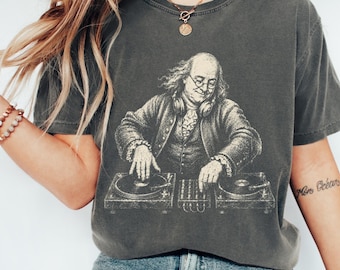 May include: A charcoal gray short-sleeve t-shirt featuring a graphic print of Benjamin Franklin wearing headphones and operating a DJ turntable setup. The shirt has a crew neckline and a relaxed fit, with the word PEPPER printed in a small circle.