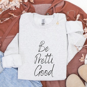 May include: A light gray sweatshirt with the words "Be Pretti Good" in black script. The sweatshirt is folded on top of a rust-colored hoodie and blue jeans. A pair of tan shoes and a necklace are also visible.