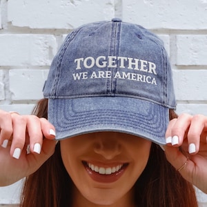 May include: A blue denim baseball cap with the text "TOGETHER WE ARE AMERICA" in white. The cap is worn by a person. The cap has a curved brim and a distressed appearance.