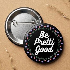 May include: A black badge with the words "Be Pretti Good" in white script, encircled by a floral wreath of purple and pink flowers. The badge has a silver pin back.