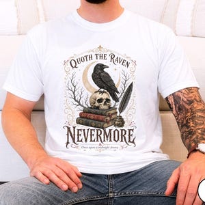 Puede incluir: Una camiseta blanca con un estampado gr&aacute;fico de un cuervo negro posado sobre un cr&aacute;neo humano, que descansa sobre una pila de tres libros. Junto a los libros hay un tintero con una pluma. El texto dice Quoth the Raven Nevermore Once upon a midnight dreary.