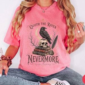 Puede incluir: Una camiseta de manga corta color rosa sand&iacute;a con un estampado gr&aacute;fico de un cuervo negro posado sobre una calavera encima de tres libros, una pluma en un tintero, una luna creciente y el texto Quoth the Raven Nevermore Once upon a midnight dreary.