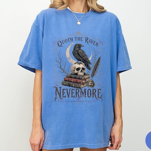 Puede incluir: Una camiseta azul de manga corta con un estampado gr&aacute;fico de un cuervo posado sobre una calavera encima de una pila de libros, con una luna creciente, una pluma y un tintero. El texto dice QUOTH THE RAVEN NEVERMORE y Once upon a midnight dreary.