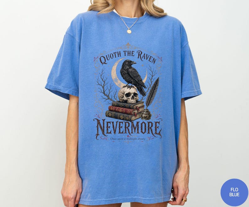 Puede incluir: Una camiseta azul de manga corta con un estampado gr&aacute;fico de un cuervo posado sobre una calavera encima de una pila de libros, con una luna creciente, una pluma y un tintero. El texto dice QUOTH THE RAVEN NEVERMORE y Once upon a midnight dreary.