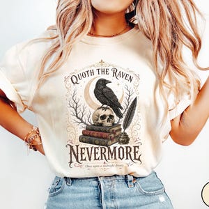 Puede incluir: Una camiseta de manga corta color marfil con un estampado gr&aacute;fico de un cuervo negro posado sobre una calavera encima de tres libros, una pluma en un tintero, ramas de &aacute;rboles desnudas, una luna creciente y el texto Quoth the Raven Nevermore.
