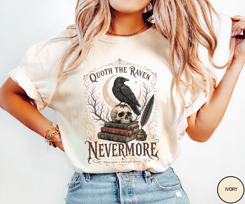Puede incluir: Una camiseta de manga corta color marfil con un estampado gr&aacute;fico de un cuervo negro posado sobre una calavera encima de tres libros, una pluma en un tintero, ramas de &aacute;rboles desnudas, una luna creciente y el texto Quoth the Raven Nevermore.