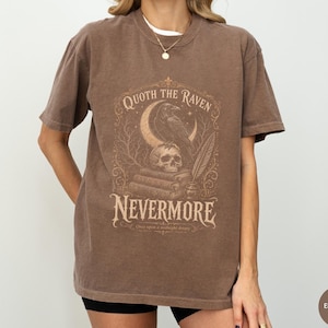 Puede incluir: Una camiseta de manga corta color marr&oacute;n espresso con un estampado gr&aacute;fico de un cuervo posado sobre una calavera encima de una pila de libros, una luna creciente, una pluma y el texto Quoth the Raven Nevermore Once upon a midnight dreary.
