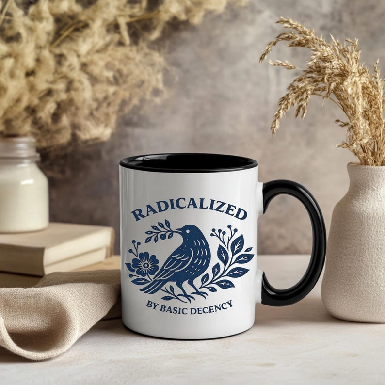 May include: White ceramic mug with a black interior and handle. The mug features a blue illustration of a bird with a branch and floral accents, and the words "RADICALIZED BY BASIC DECENCY" in blue lettering.