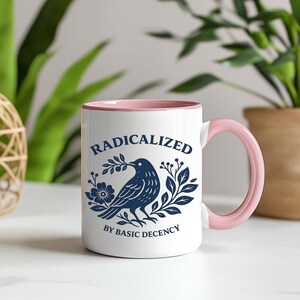 May include: White ceramic mug with a pink interior and handle. The mug features a navy blue illustration of a bird with a branch in its beak, surrounded by floral elements. The text "RADICALIZED BY BASIC DECENCY" is printed on the mug.