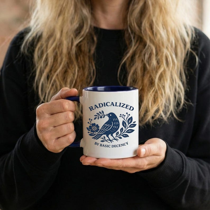 May include: White ceramic mug with a blue interior and handle. The mug features the word "RADICALIZED" above a blue illustration of a bird with a floral and leaf design. The text "BY BASIC DECENCY" is below the bird.