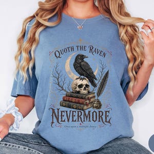 Puede incluir: Una camiseta de color azul vaquero con un estampado gr&aacute;fico de un cuervo negro posado sobre un cr&aacute;neo humano encima de una pila de tres libros. Junto a los libros hay un tintero con una pluma. El texto dice Quoth the Raven, Nevermore, Once upon a midnight dreary.