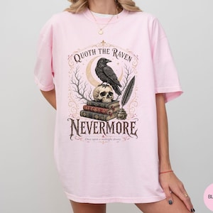 Puede incluir: Una camiseta rosa claro con un estampado gr&aacute;fico de un cuervo negro posado sobre una calavera encima de una pila de libros, con una pluma en un tintero. El texto dice Quoth the Raven, Nevermore y Once upon a midnight dreary en letras oscuras.
