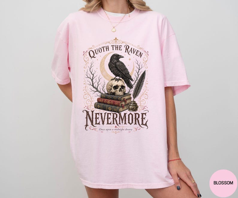Puede incluir: Una camiseta rosa claro con un estampado gr&aacute;fico de un cuervo negro posado sobre una calavera encima de una pila de libros, con una pluma en un tintero. El texto dice Quoth the Raven, Nevermore y Once upon a midnight dreary en letras oscuras.