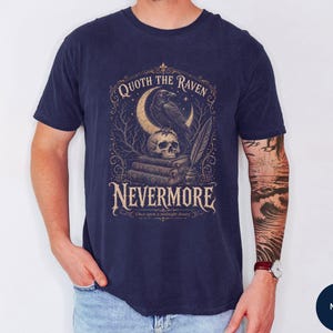 Puede incluir: Una camiseta azul marino con un estampado gr&aacute;fico de un cuervo posado sobre una calavera encima de una pila de libros, con una luna creciente, una pluma y el texto "QUOTH THE RAVEN NEVERMORE" y "Once upon a midnight dreary" en fuente beige.