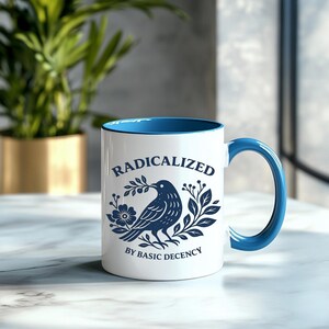 May include: White ceramic mug with a blue interior and handle. The mug features a navy blue illustration of a bird holding a branch, surrounded by floral elements. Text on the mug reads "RADICALIZED BY BASIC DECENCY."