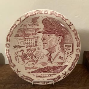May include: A decorative plate with a maroon and white design. The plate features a portrait of General Douglas MacArthur, along with text that reads "Graduated from Westpoint-1903" and "The Island Fortress of Corregidor".