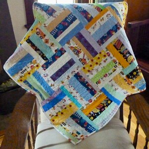 May include: A patchwork quilt with a white background and colorful stripes in shades of blue, yellow, green, and purple. The quilt features a variety of small, colorful patterns, including trucks, flowers, and geometric shapes.