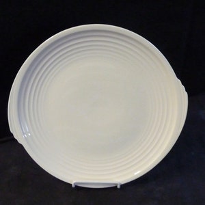 May include: A white, round serving platter with a ridged design. The plate has two small handles and is displayed on a white stand. The plate's design features concentric circles, adding a touch of elegance to the tableware.