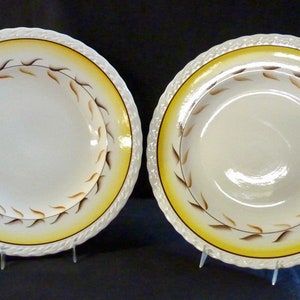 May include: Two white ceramic bowls with a yellow and brown leaf design. The bowls have a raised rim and are on display stands.