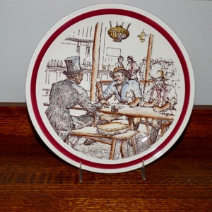 May include: A white ceramic plate with a red rim features a vintage illustration of men sitting at a table in a saloon. The scene includes a bar, a chandelier, and several patrons.