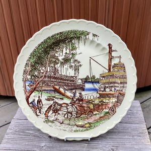 May include: A white ceramic dinner plate with a scalloped edge features a vintage illustration of two paddlewheel steamboats on a river. The scene includes trees, a dock, and people in period clothing.