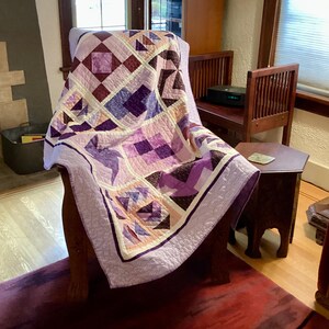 May include: A purple, pink, and brown patchwork quilt with a geometric design is draped over a wooden armchair. The quilt is made with various fabrics and patterns, including squares, triangles, and diamonds.